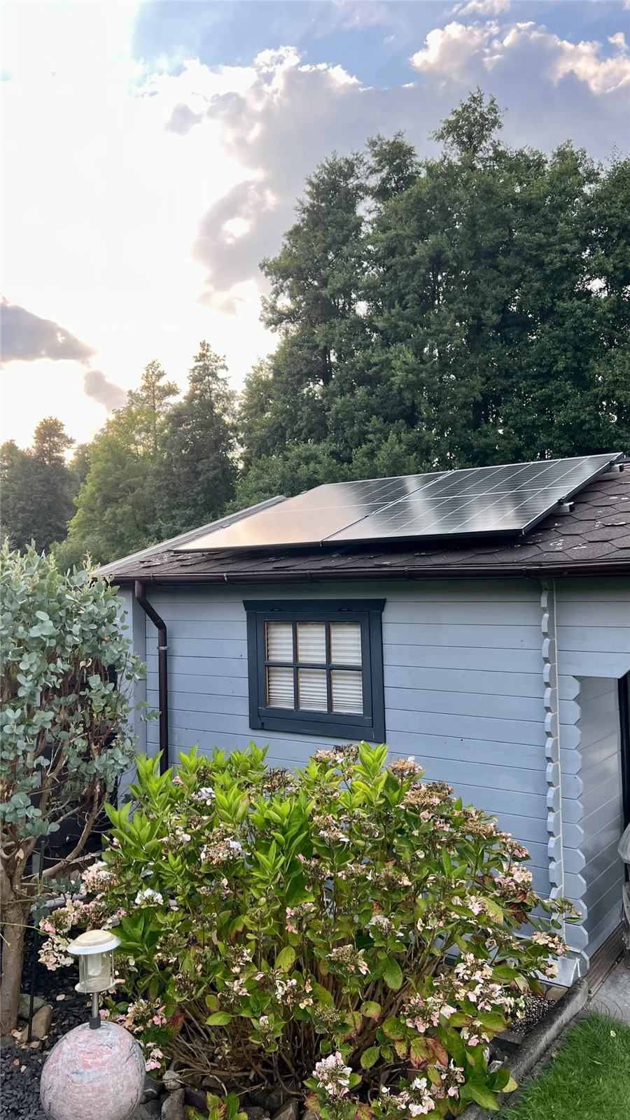 Von M-Energie installierte Solarmodule auf dem Dach eines hellblauen Gartenhauses zwischen Bäumen und Hortensien